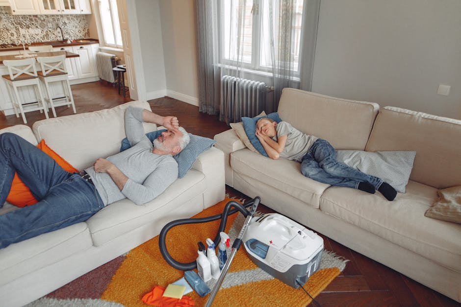 A person wearing a light grey hoodie is adjusting a decorative cushion on a grey upholstered sofa in a well-lit living room. The cushion has a black and white checkered pattern, complementing two othe