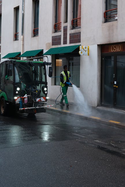 A worker from Kentish Town Carpet Cleaners is engaged in surface cleaning on a city street, using a high-pressure water jet to clean the asphalt pavement in front of a commercial building. The worker 