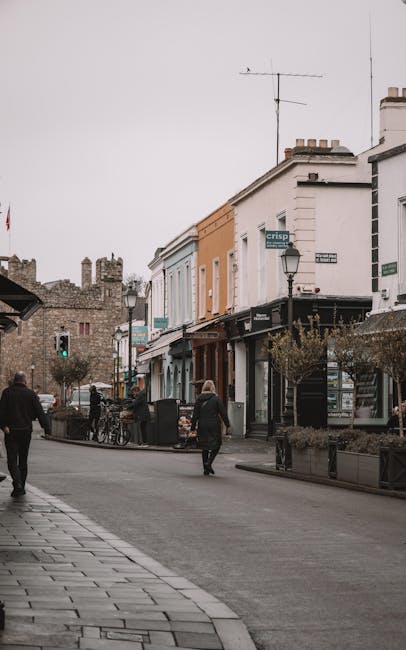 A street scene on Kentish Town Road featuring a row of shops with colorful facades, including a light blue and orange building, under an overcast sky. Pedestrians are walking along the paved sidewalk, which is lined with potted plants and a black metal fence. The shops have large glass windows, with some displaying signage, and a lamppost is visible on the corner. In the background, older brick buildings and a television antenna on a rooftop can be seen. The overall atmosphere is calm, with a focus on the commercial area that could benefit from professional cleaning and surface maintenance, as provided by Kentish Town Carpet Cleaners.