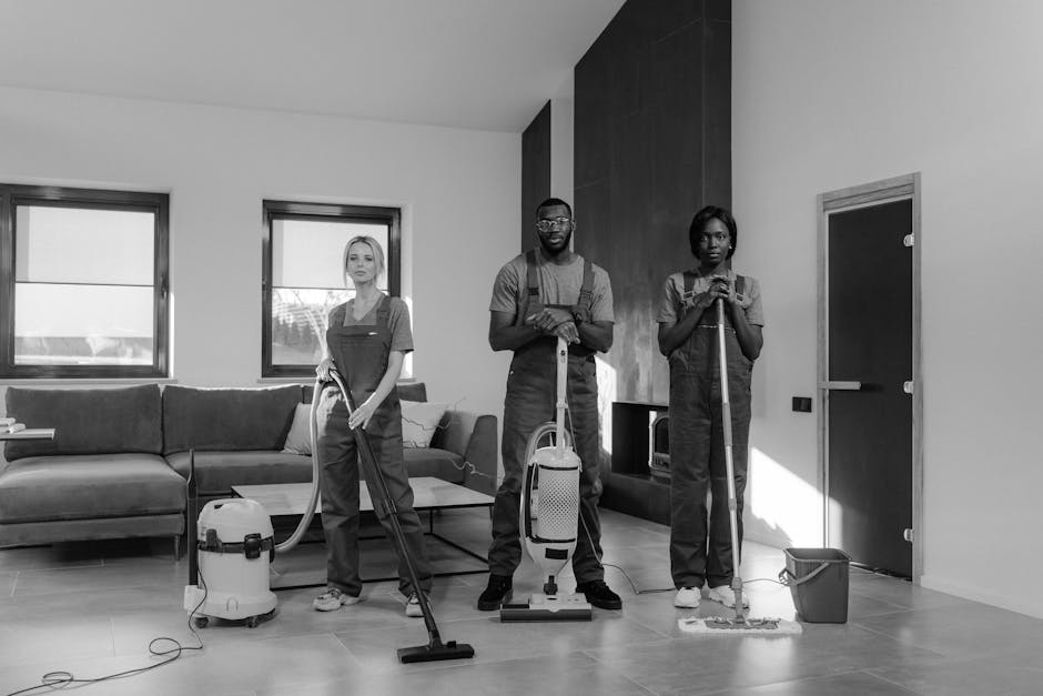 Three professional cleaners equipped with vacuum, mop, and cleaning cloths standing in a modern living room with a dark-colored sofa, two large windows, and a wooden accent wall. The room features til